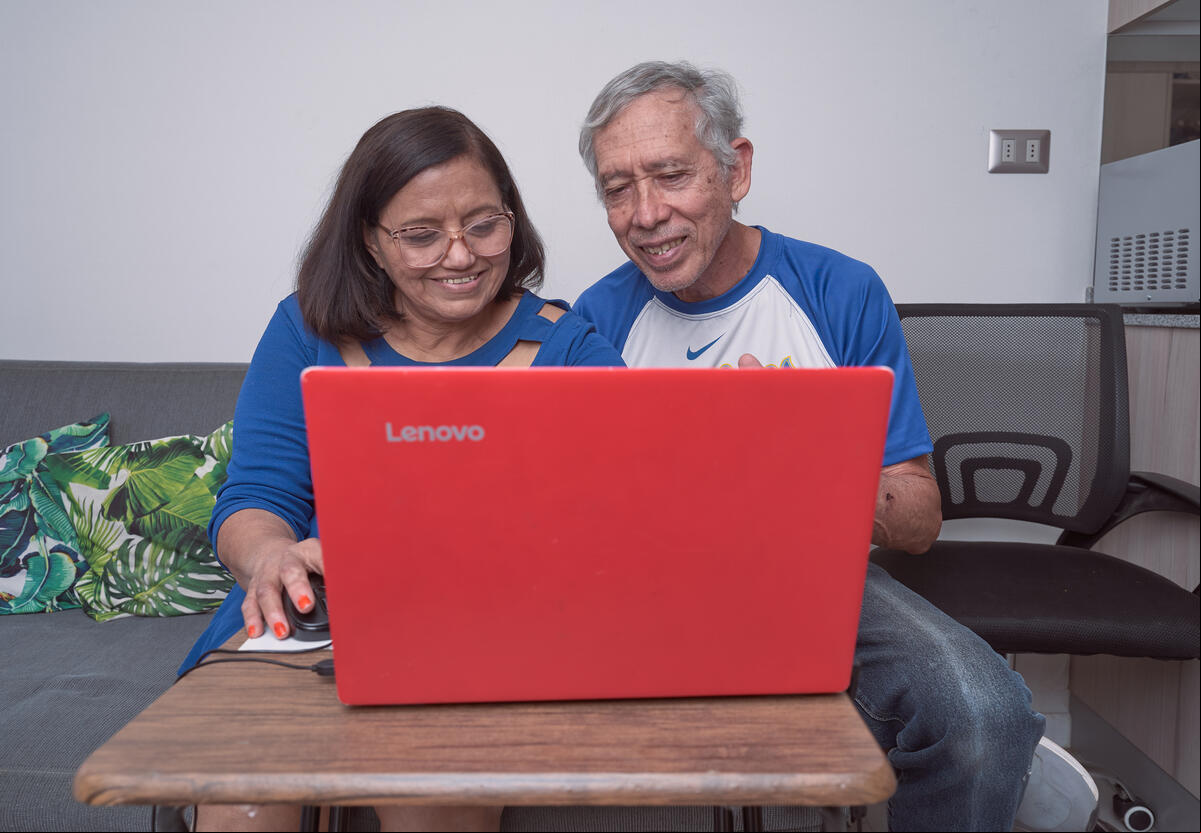 Un hombre y una mujer mayores, en un sofá, ven un computador que está frente suyo, sobre una pequeña mesa