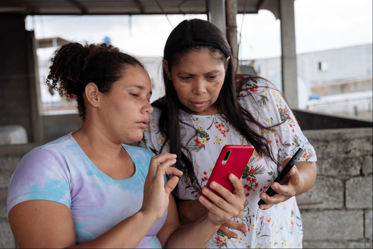 Dos mujeres. Una de ellas sostienen un teléfono móvil y se lo muestra a la otra.